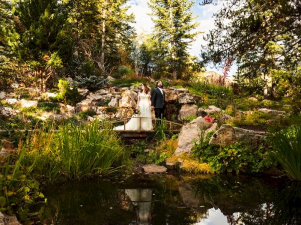 Bride and groom pose outdoors beside a pond, on a small bridge, with tall trees and rocks in a garden.