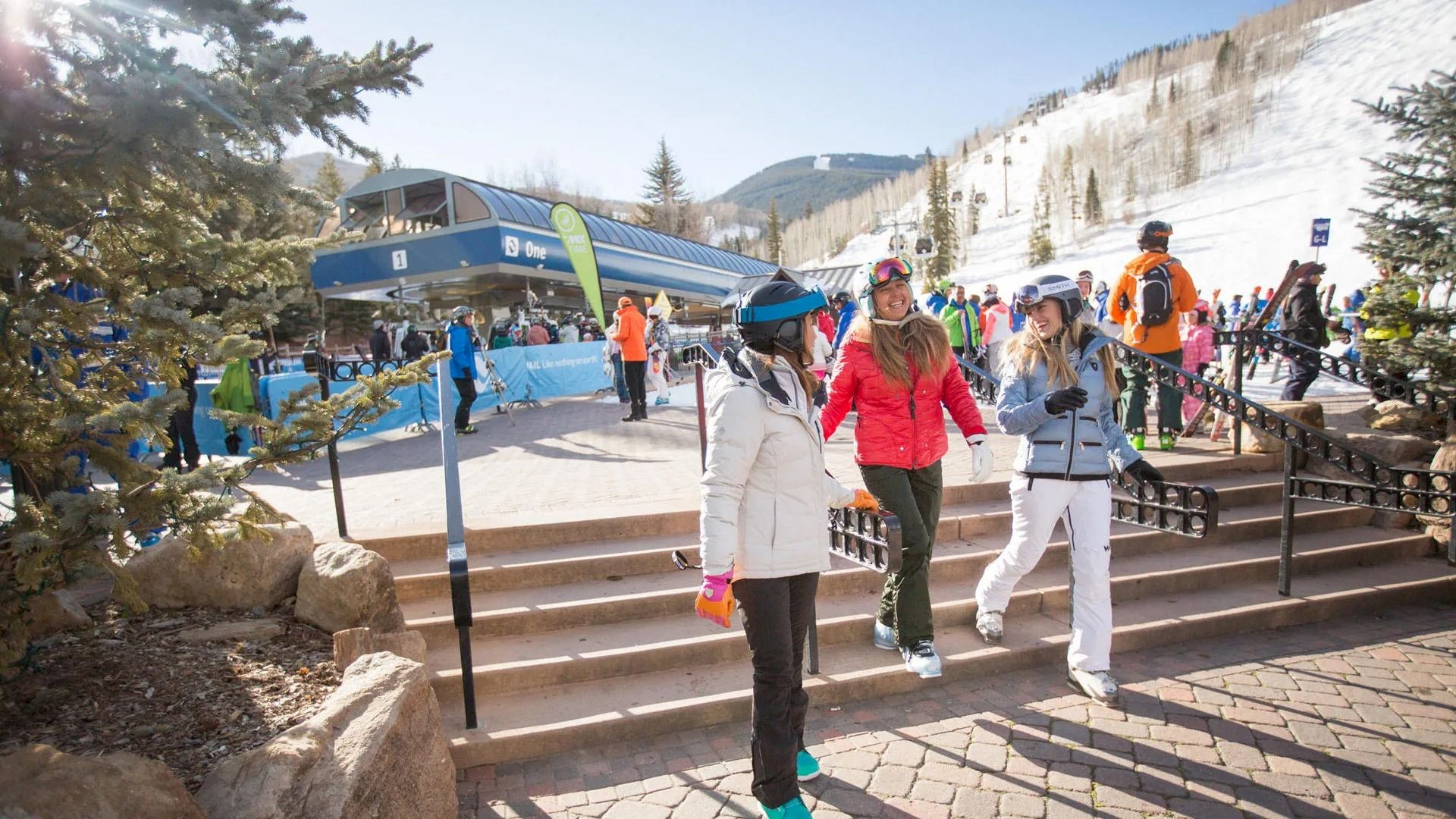 People in winter clothing at a ski resort, walking near a ski lift station with snow-covered mountains and trees in the background.