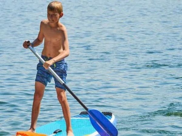 A shirtless boy is standing on a paddleboard in the water, using a paddle to navigate on a sunny day.