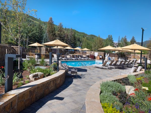 A serene outdoor pool area is surrounded by lounge chairs, umbrellas, and lush greenery against a backdrop of forested hills under a clear blue sky.