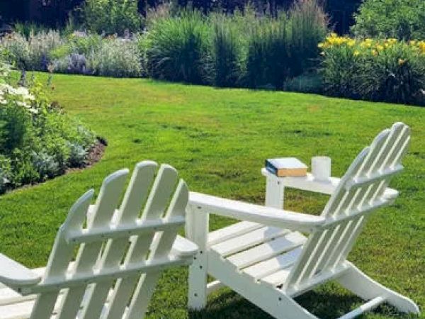 Two white Adirondack chairs sit on a green lawn, facing a garden. A small table between them holds a book and a white cup, hinting at relaxation.