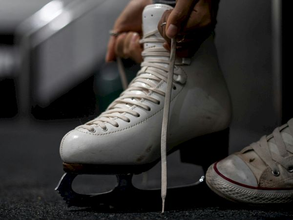 A person is tying the laces of a white ice skate on their foot, with the other foot wearing a white sneaker.