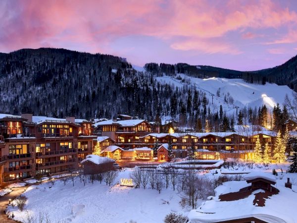A snowy mountain ski resort at dusk, with a lit hotel village and ski slopes winding through the valley.