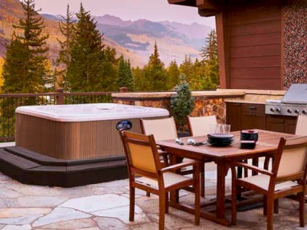 The image shows an outdoor patio with a hot tub, dining table, chairs, and a grill, surrounded by a scenic mountainous forest landscape.
